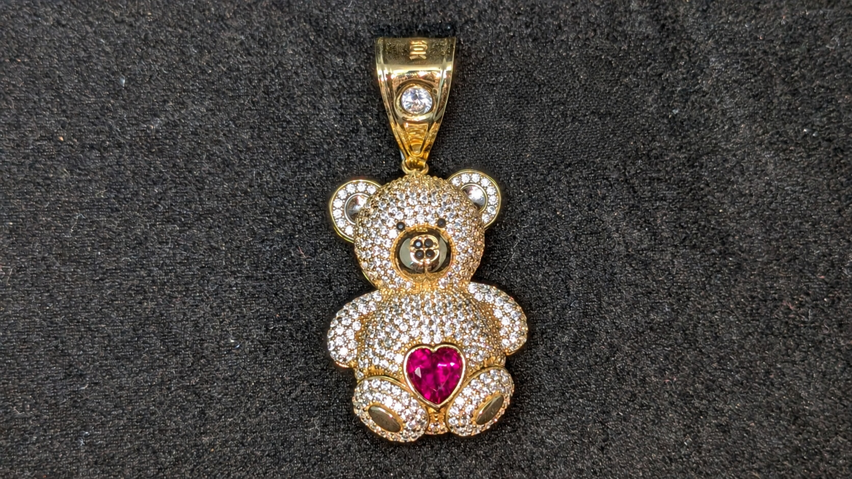 Teddy bear pendant with clear stone accents and a pink heart center on a dark jewelry display
